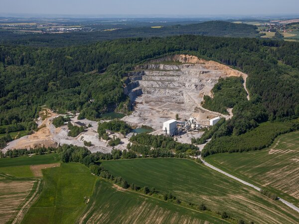 Luftaufnahme eines großflächigen Steinbruchs am Berghang, umgeben von Wald und Feldern